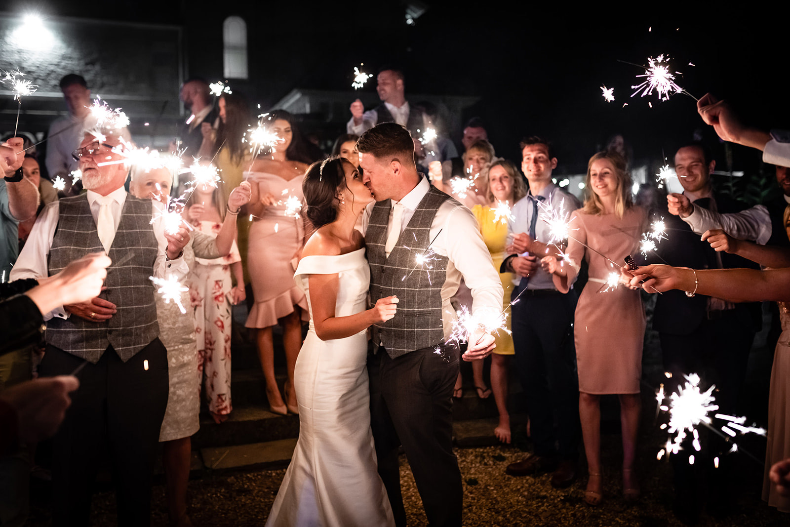 A bridal couple kissing surrounded by guests with sparklers at night